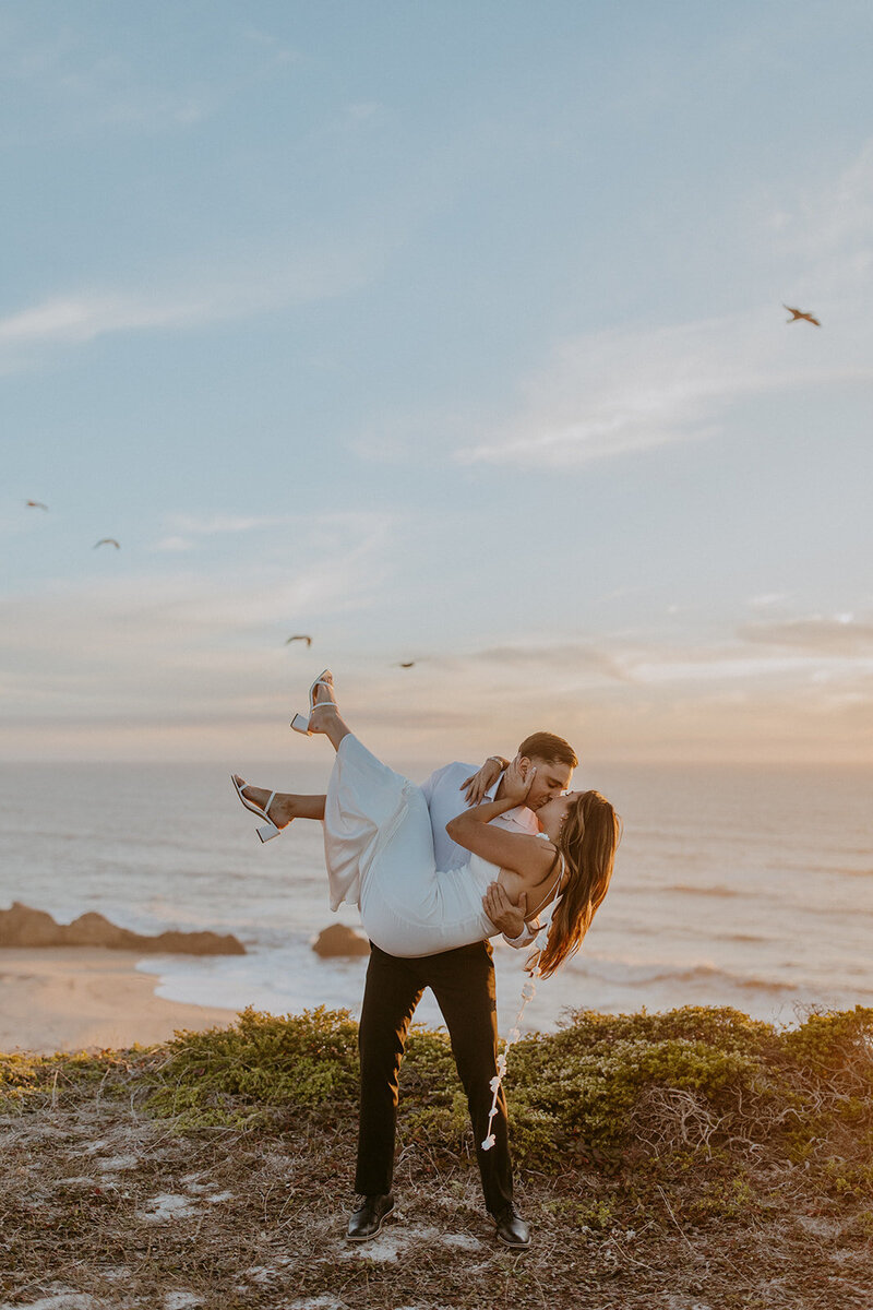 couple kisses on a cliff during an engagement session at half moon bay in california