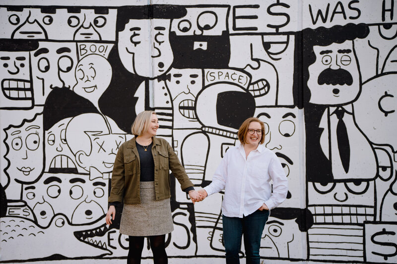 A couple smiling and holding hands while standing in front of a wall with a black and white mural painted on it.
