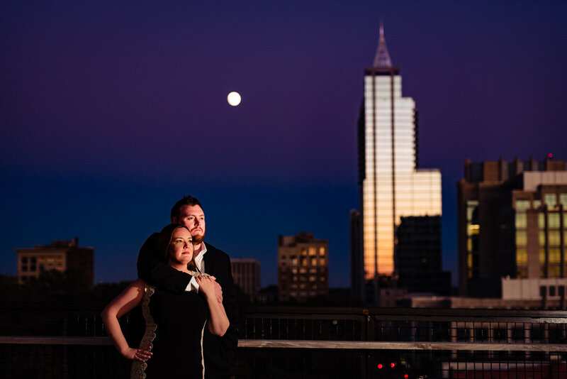 Well-dressed couple stands on a balcony overlooking the downtown Raleigh skyline during their engagement photo session