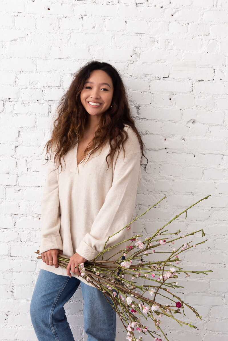 Floral designer holds branches and smiles standing against white brick wall