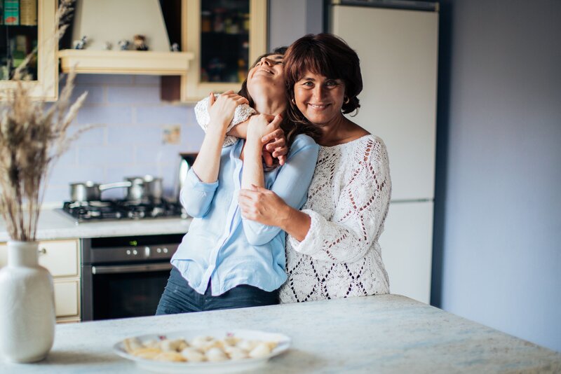 mother hugs her adult daughter and smiles in a kitchen