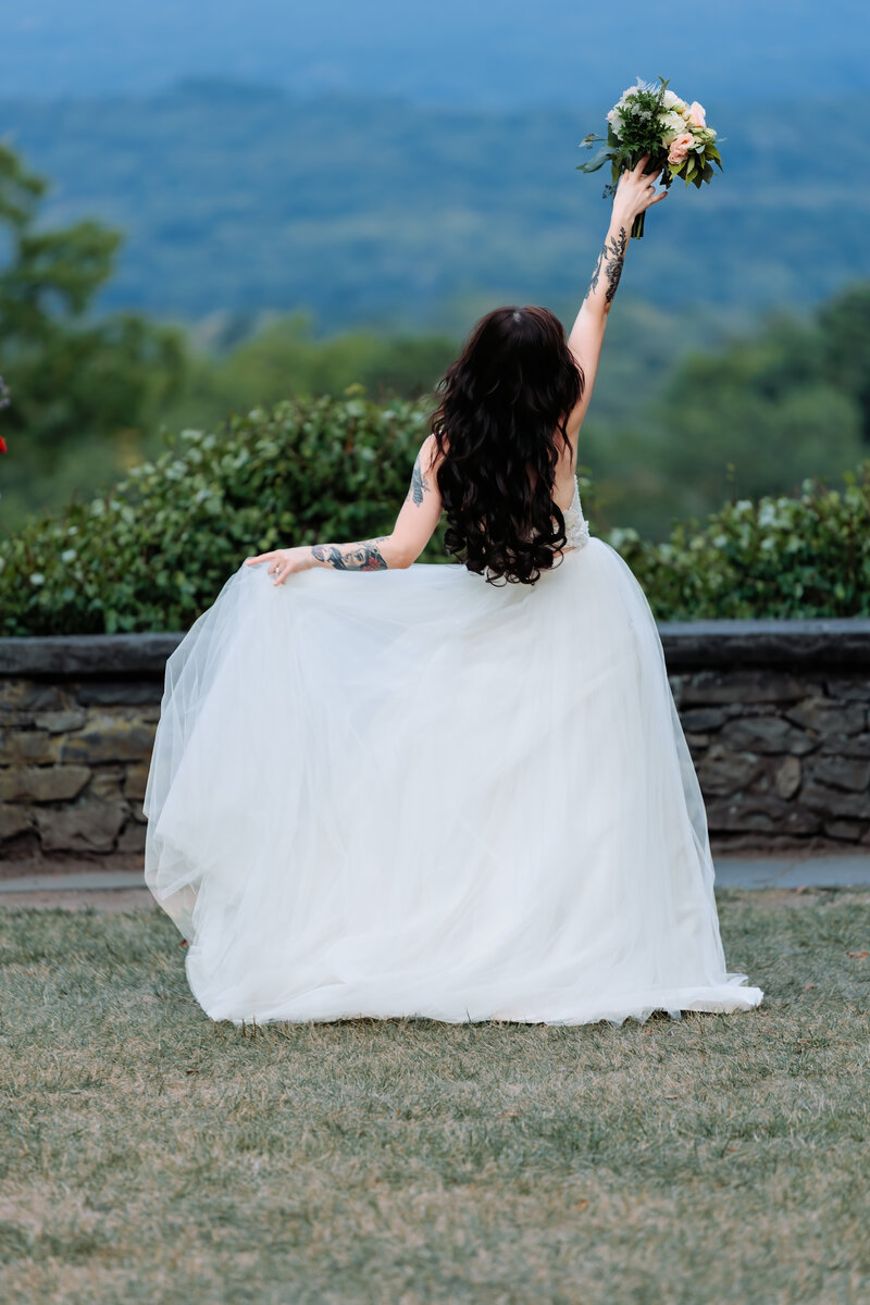 Dark haired, Tattooed Bride celebrates her wedding by holding her bouquet high in the air and looking out over the view at Grey Towers Historic Site in Milford, Pennsylvania