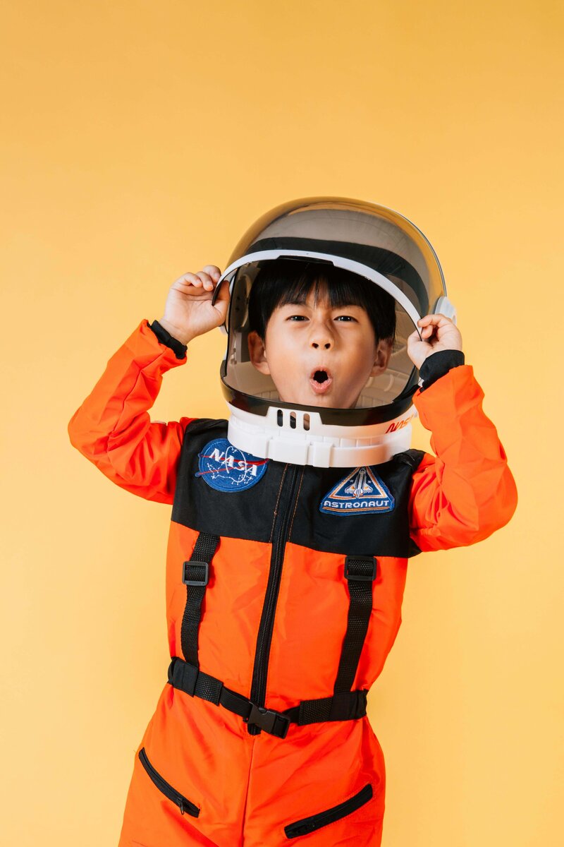 A young child in an orange astronaut costume excitedly holding up their helmet, standing against a vibrant yellow background.