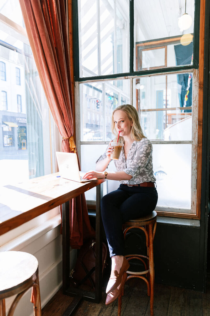 Website designer, Sarah Blodgett, sits on a wooden stool drinking iced coffee in a cafe at her laptop