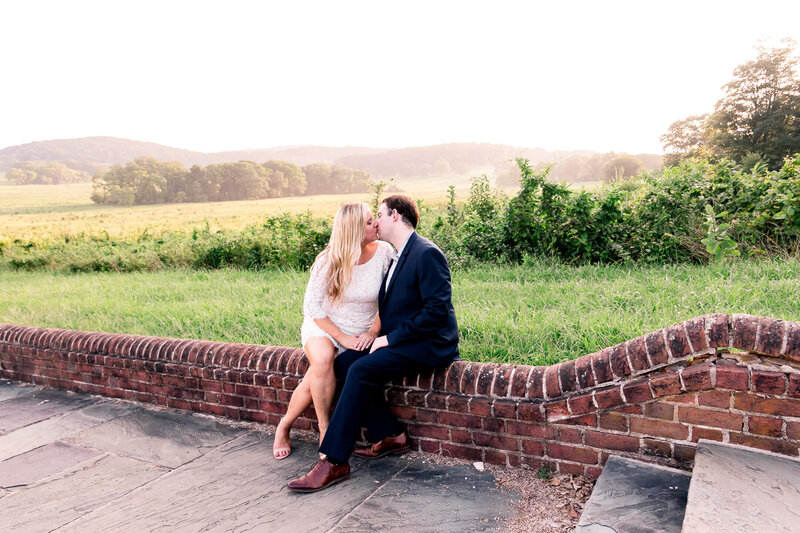 Couple kissing as they sit on a brick wall over looking open fields and hills