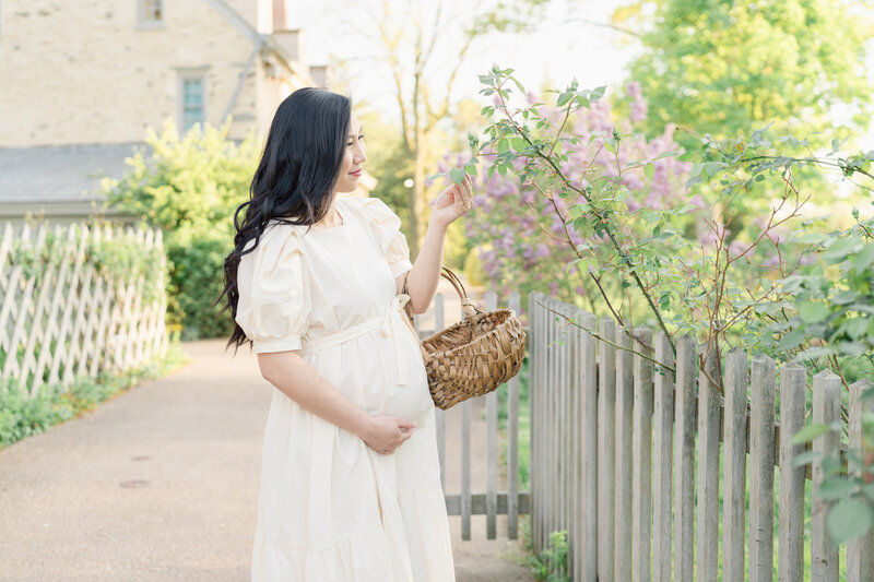 Pregnant woman wearing a cottagecore maternity dress holds a gathering basket and looks at the garden.