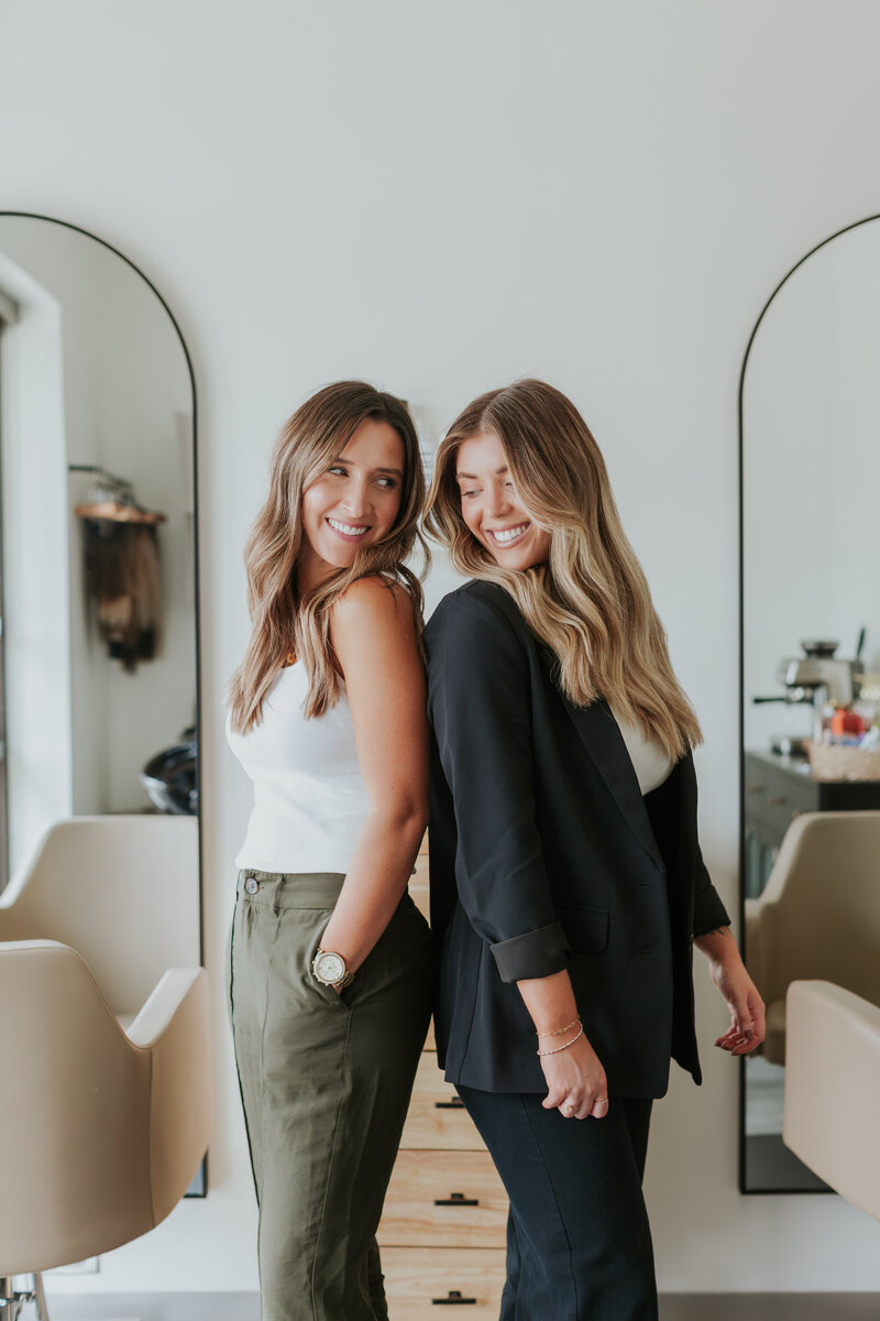 Team photo of Bekah Tutwiler and Kendal Harler Fadale, owners of Aurora South Salon, smiling and standing back to back in the salon