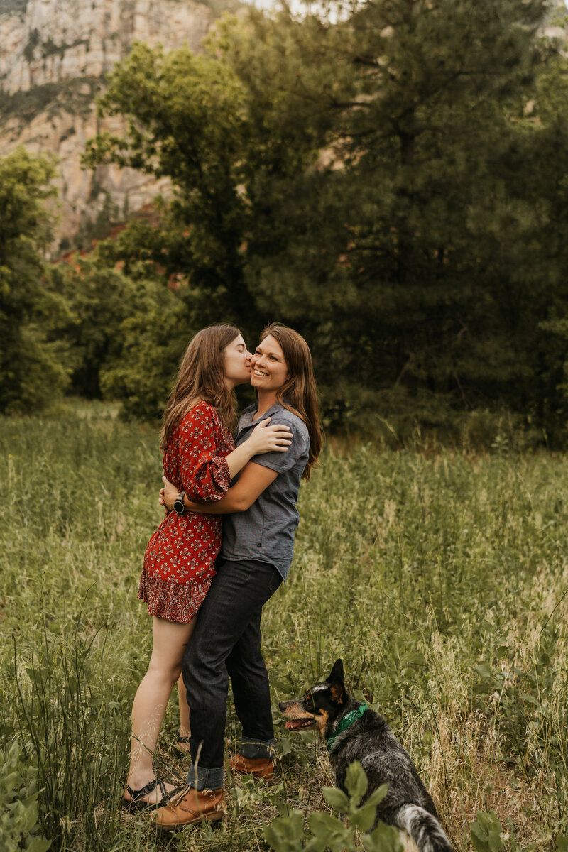 A person kissing their partner on the cheek with their dog at their feet 