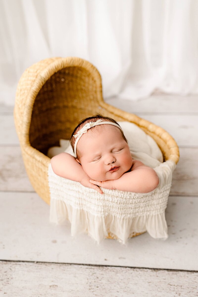 newborn baby girl is laying with her arms out and her hands underneath her cheek and a Moses Basket in a studio newborn session