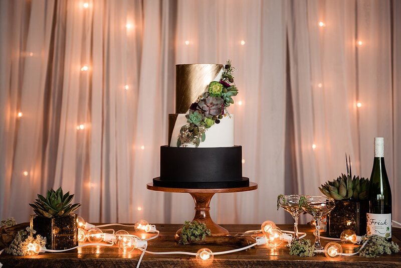 Gold, ivory and black 3 tier wedding cake with succulents, string lights and champagne