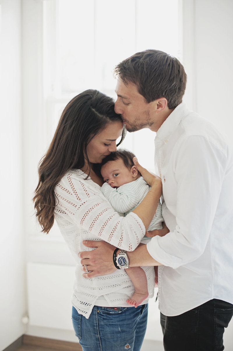 A couple kiss as they hold their 4 month old baby boy during an at home photo shoot in Kensington