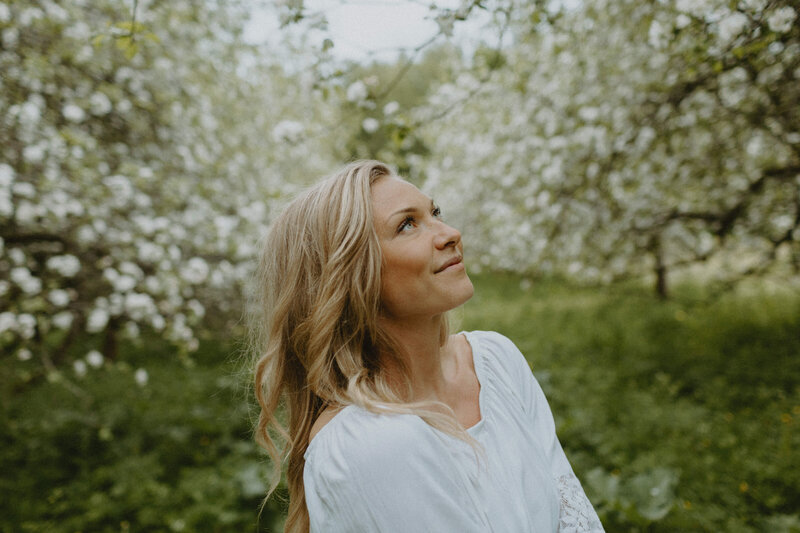 Muotokuva naisesta, joka seisoo kukkivien omenapuiden keskellä ja katsoo hymyillen ylöspäin Helsingissä Malkartanon Omenatarhassa