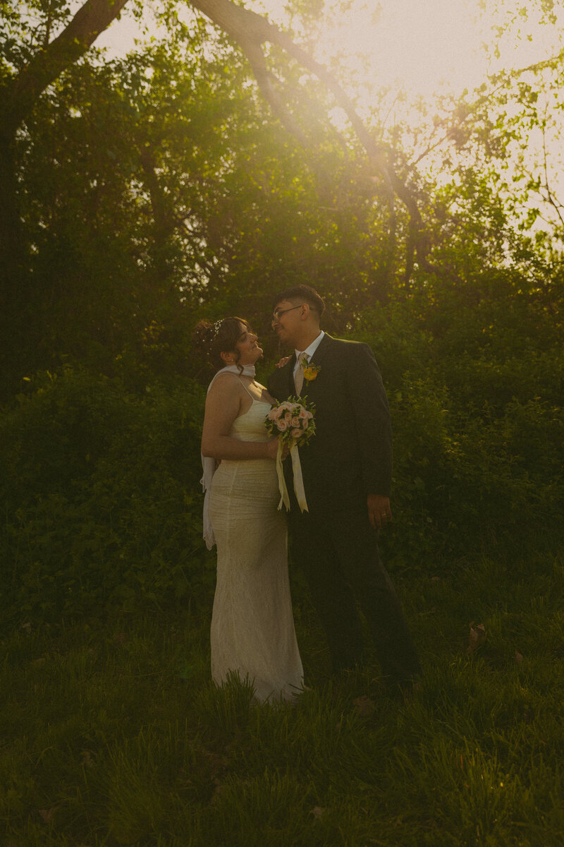 Newlyweds gaze into eachothers eyes on a brisk summer evening while the sun glares between their faces and her veil gets carried in the wind