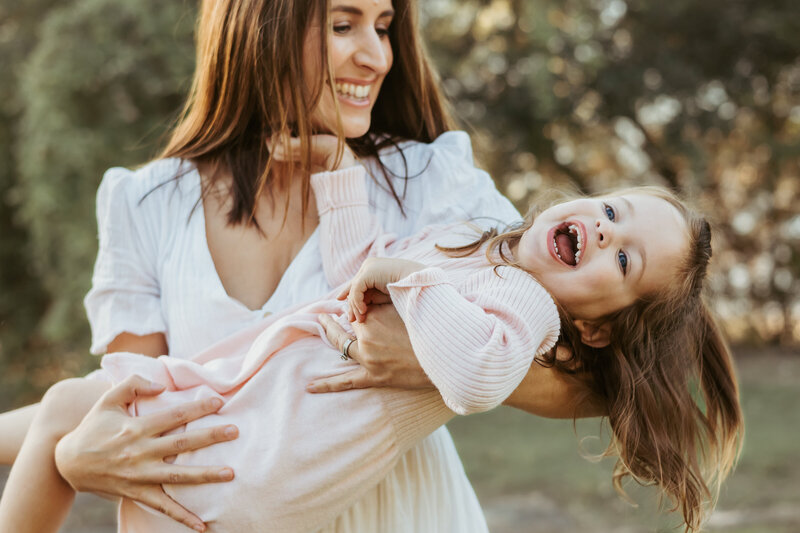 Mum with brown hair in a white dress swinging around her three year old daughter in a pink dress and smiling widely.