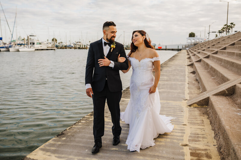 Newly married husband and wife walk along a wooden pier while arm in arm and smiling