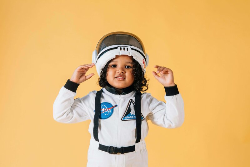 A young child dressed in a white astronaut costume with NASA patches and a helmet is smiling and gesturing with their hands against a yellow background.