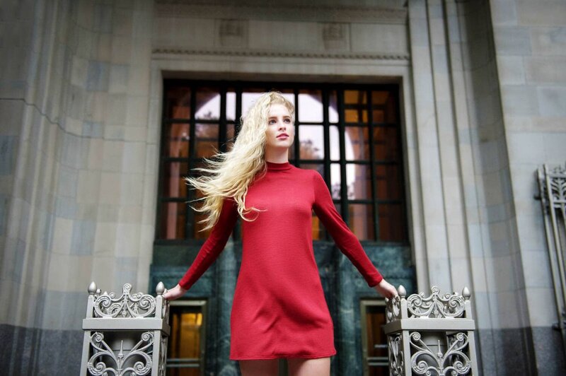 Blonde haired young lady in a long sleeve red dress posed in front of New York building.