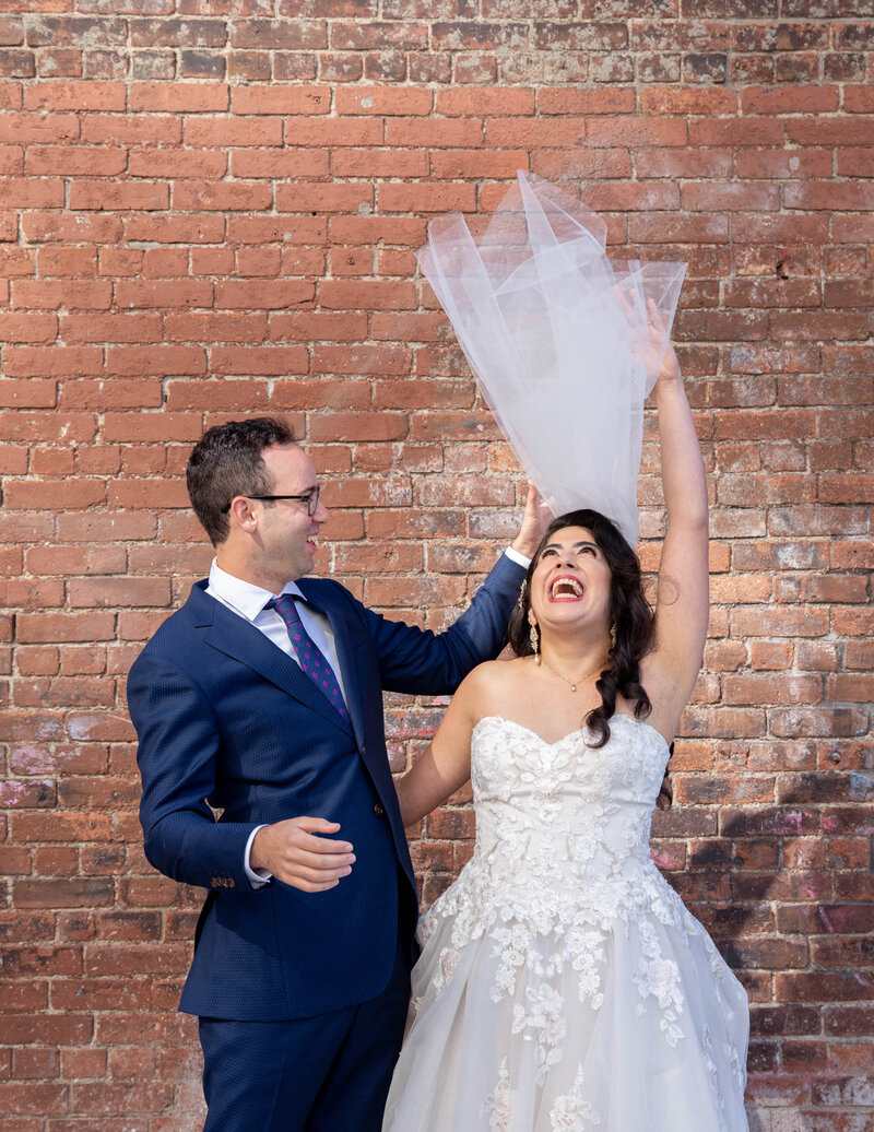 A bride flipping her veil over her head as her groom smiles next to her.