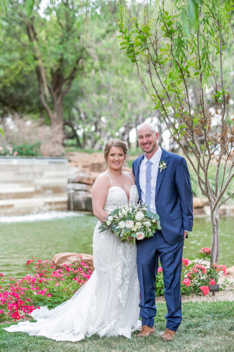 Bride wearing a white  lace  wedding dress with white and green bouquet standing with groom wearing a blue suit in front of pink flowers and a garden waterfall