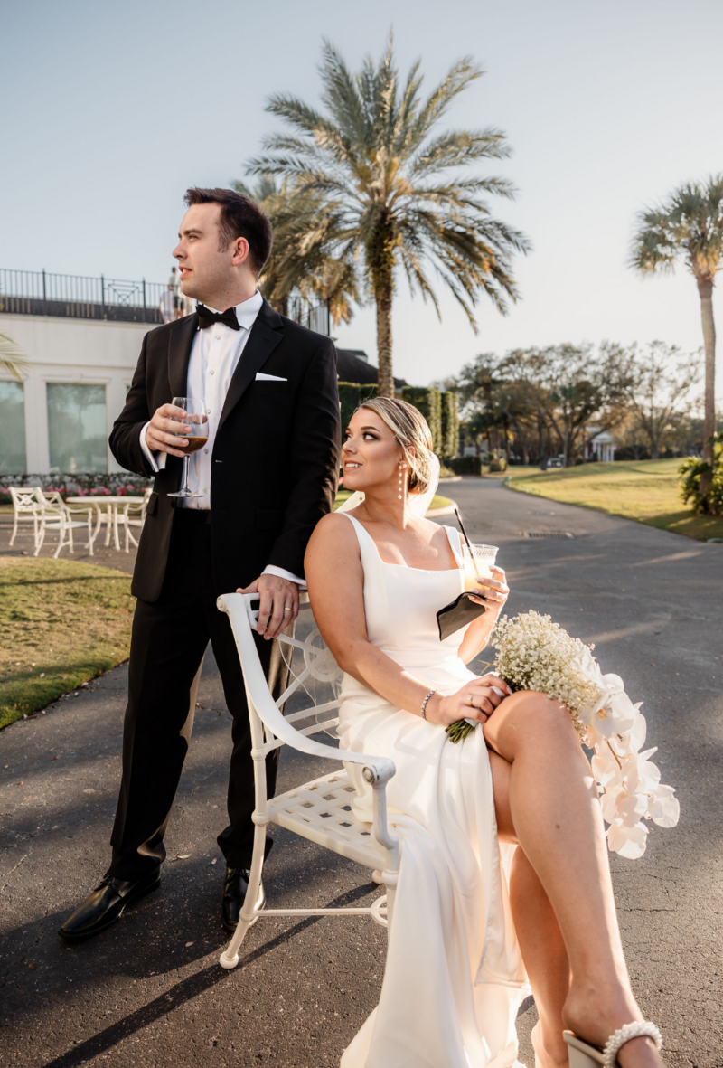 Bride in wedding dress with white flower bouquet sitting in chair looking up a groom standing wearing a black tux