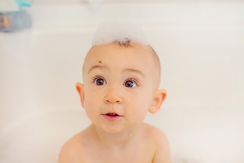 Baby is sitting in the bathtub, looking up, with bubbles on her head.