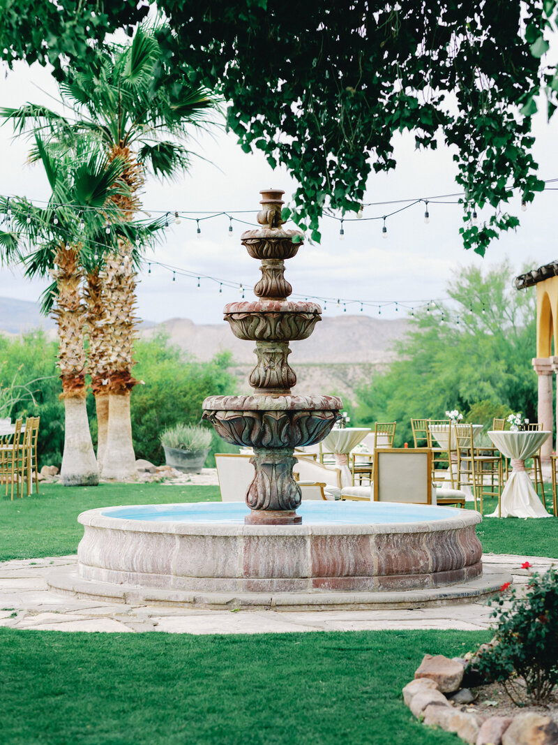 A stone fountain in a lush outdoor setting, surrounded by palm trees and event tables, capturing the elegance of Socially Significant Events’ luxury event design.