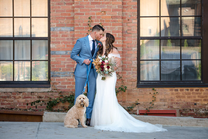 Couple-standing-and-kissing-for-a-portrait-with-their-dog-in-front-of-a-brick-wall-at-The-Foundry-Hotel-in-Asheville