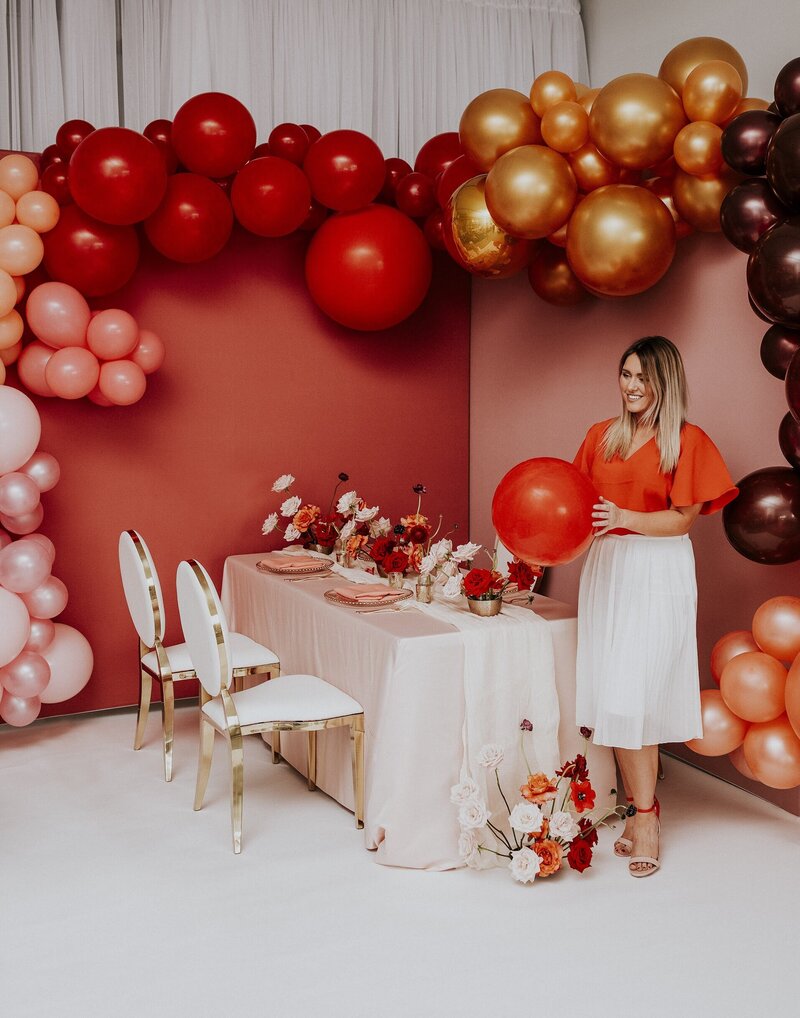 Brittany Frid is wearing a cherry red and white outfit while holding a red balloon, standing beside a modern glamorous guest table with red and blush florals set up in front of a large two-panelled red and pink backdrops covered across the top with a huge balloon garland with red, gold and burgundy balloons in Ottawa Ontario.