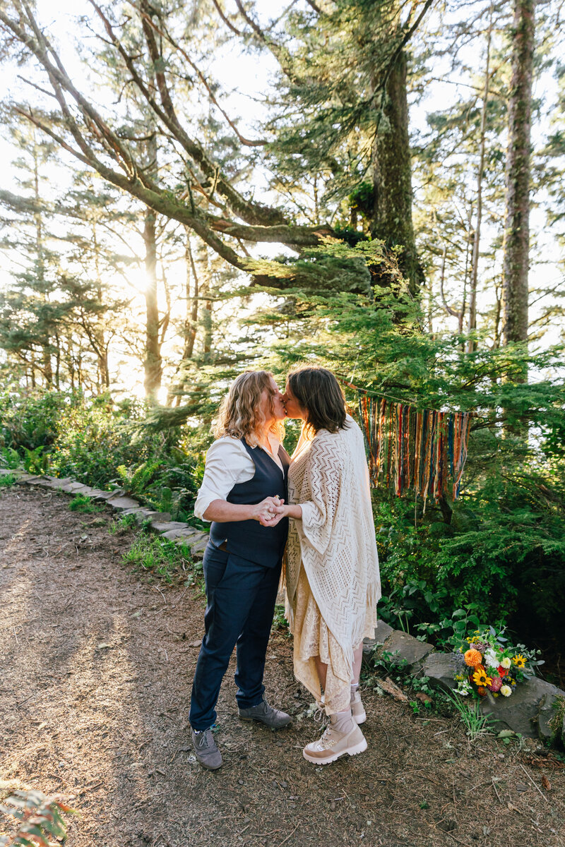 Two brides, one wearing a navy vest and slacks, while the other has a lacy cream poncho and hiking boots, embrace in a kiss while holding hands as the sun shines through the trees and their rainbow backdrop along the Cape Perpetua Lookout on the Oregon Coast during their adventure elopement. | Erica Swantek Photography