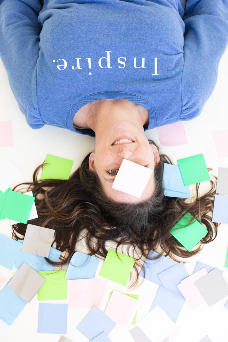 social media strategist and course creator is lying on the white floor and all around her are sticky notes in blue-green and pastel gray. her face is covered by a white paper and she is sleeping in a blue  coat