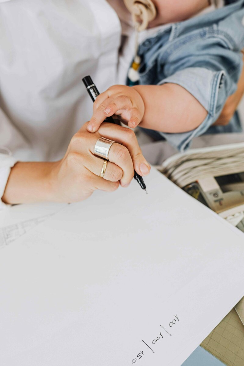Working mom helping a baby hold a pen, both hands resting on a blank sheet of paper with some numbers written in the corner. The baby is wearing a denim jacket and sitting on the adult’s lap.


