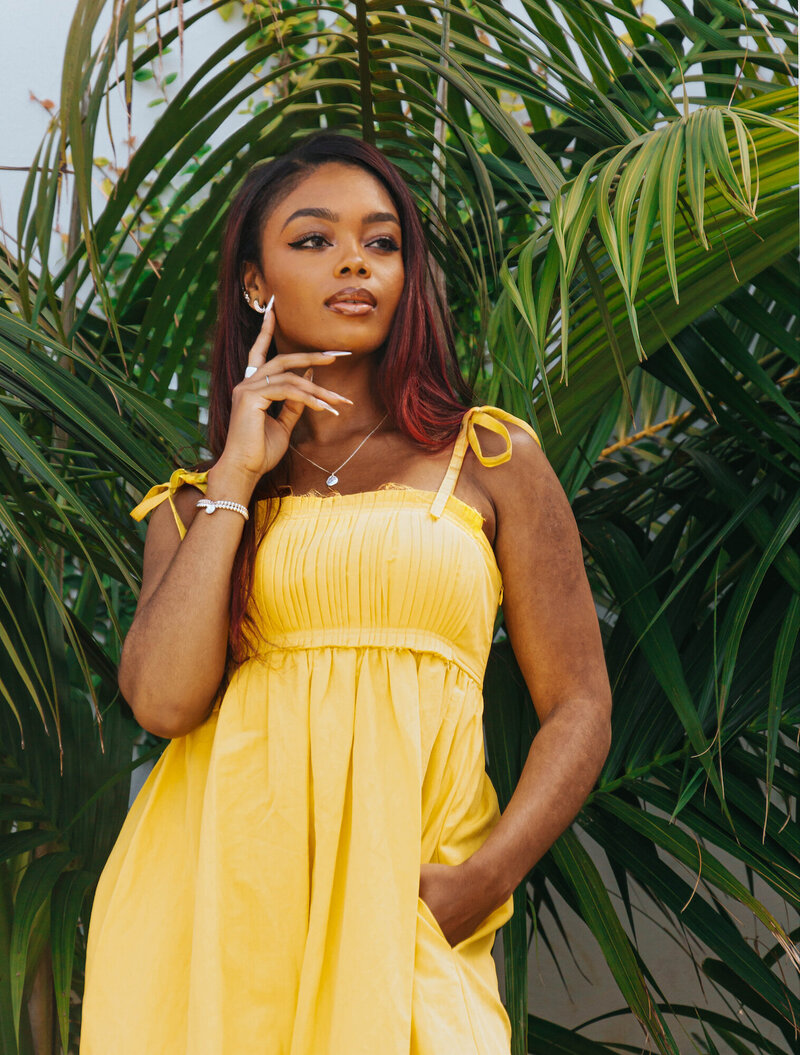 Tahirah standing in front of a palm tree in a yellow dress, fingers gently touching her chin and jaw