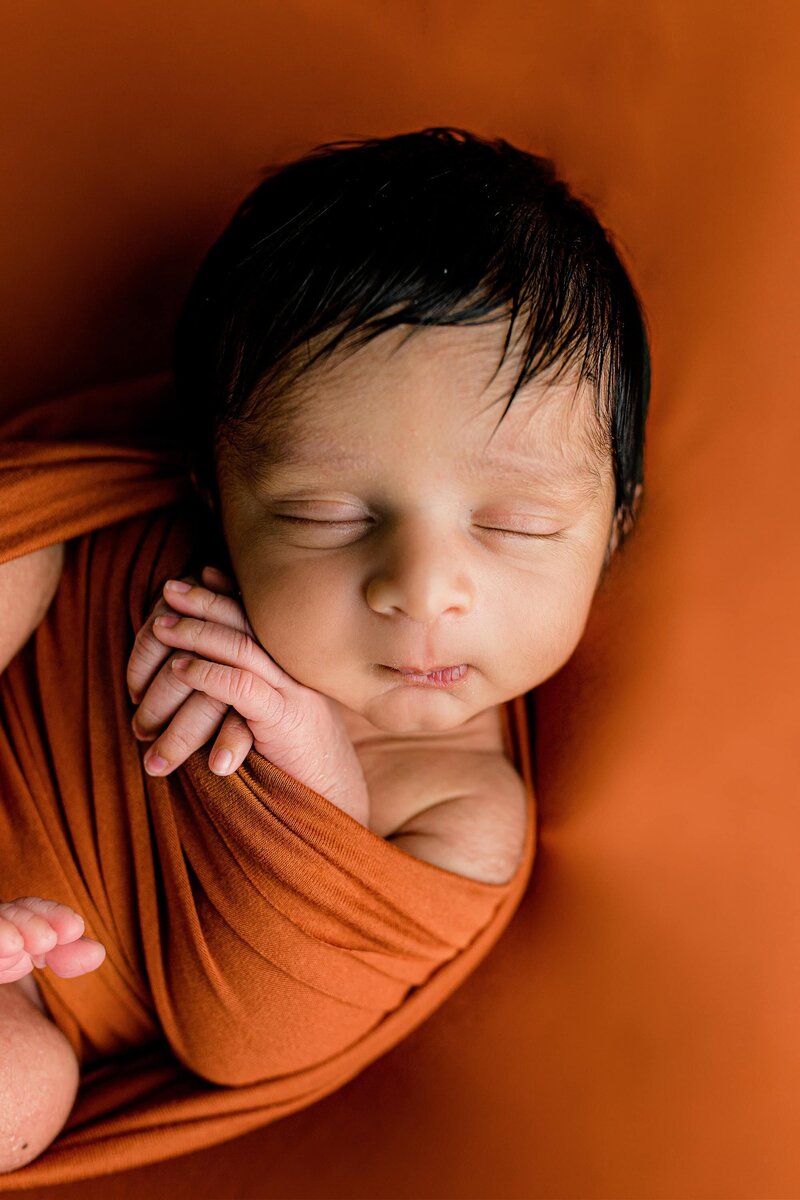 Close-up image of a baby's features with his hands up by his face where you can see his dark hair and amazing eyelashes as he's wrapped in a rust colored background during his Charlotte Studio newborn session