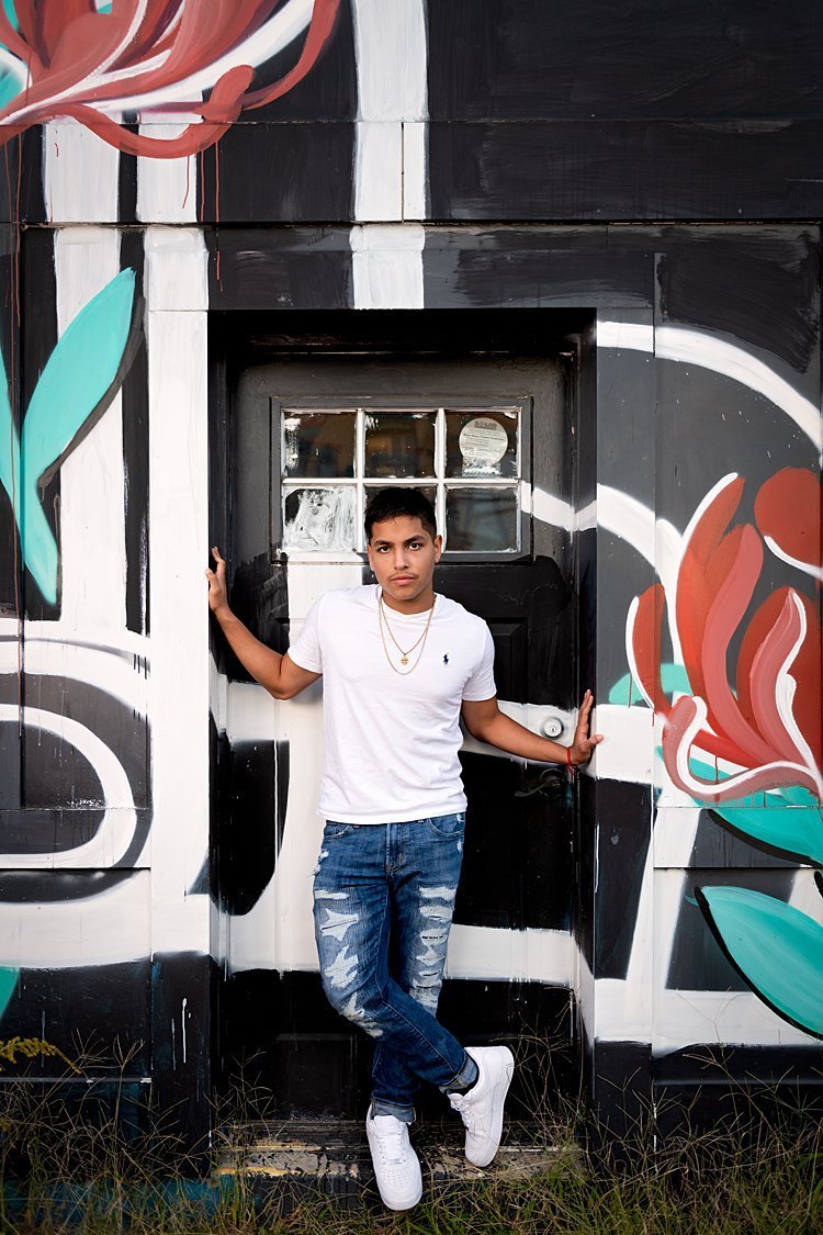 High school senior boy standing in doorway of building painted with a mural of black, white and red roses in Pittsburgh, PA