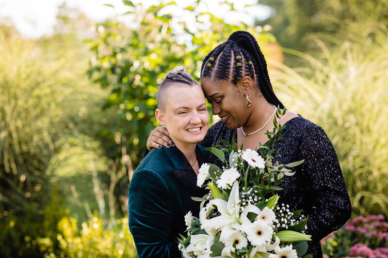 Two brides share an intimate moment surrounded by greenery, one holding a bouquet