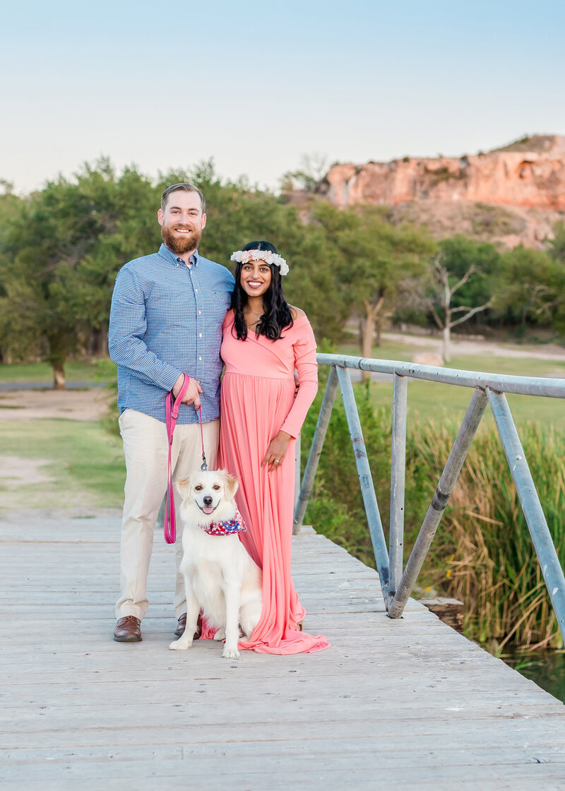 Man and woman posed with white dog standing on a bridge with trees and rocks in the background.  Woman is wearing a pink maternity dress with a white flower crown and man is wearing a blue shirt and white khakis.