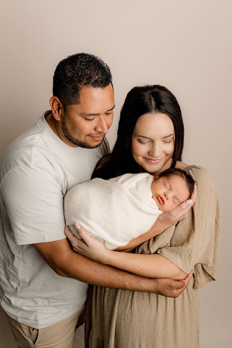 Mother cradles her newborn son as he is wrapped in a cream-colored wrap she is wearing a brown dress and dad is standing beside her in a neutral colored top