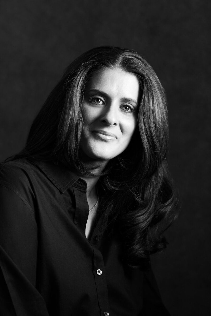 Black & White photo. Woman in black shirt looking at camera against black background.