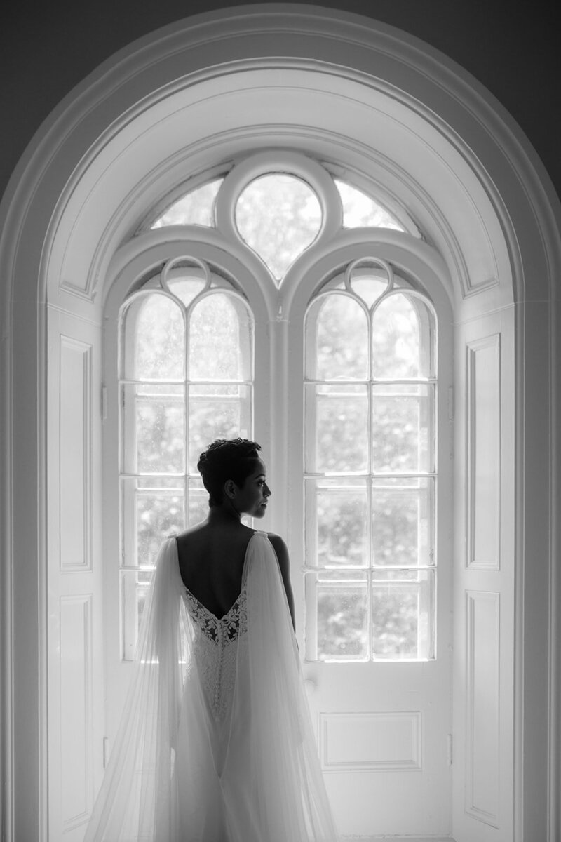 A bride in front of a grand arched window, backlit in a monochrome setting.