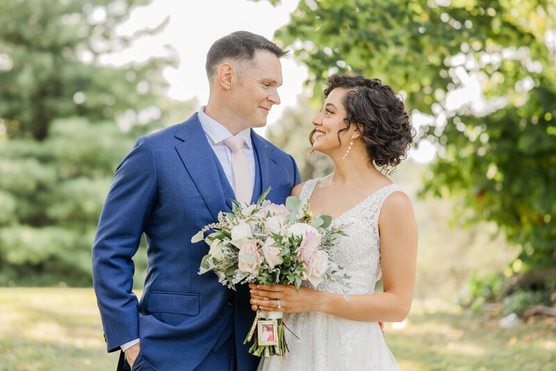 A newlywed couple, the man in a blue suit and the woman in a white lace dress, smiling at each other while holding a bouquet in a sunny park, perfectly capturing the essence of Iowa weddings