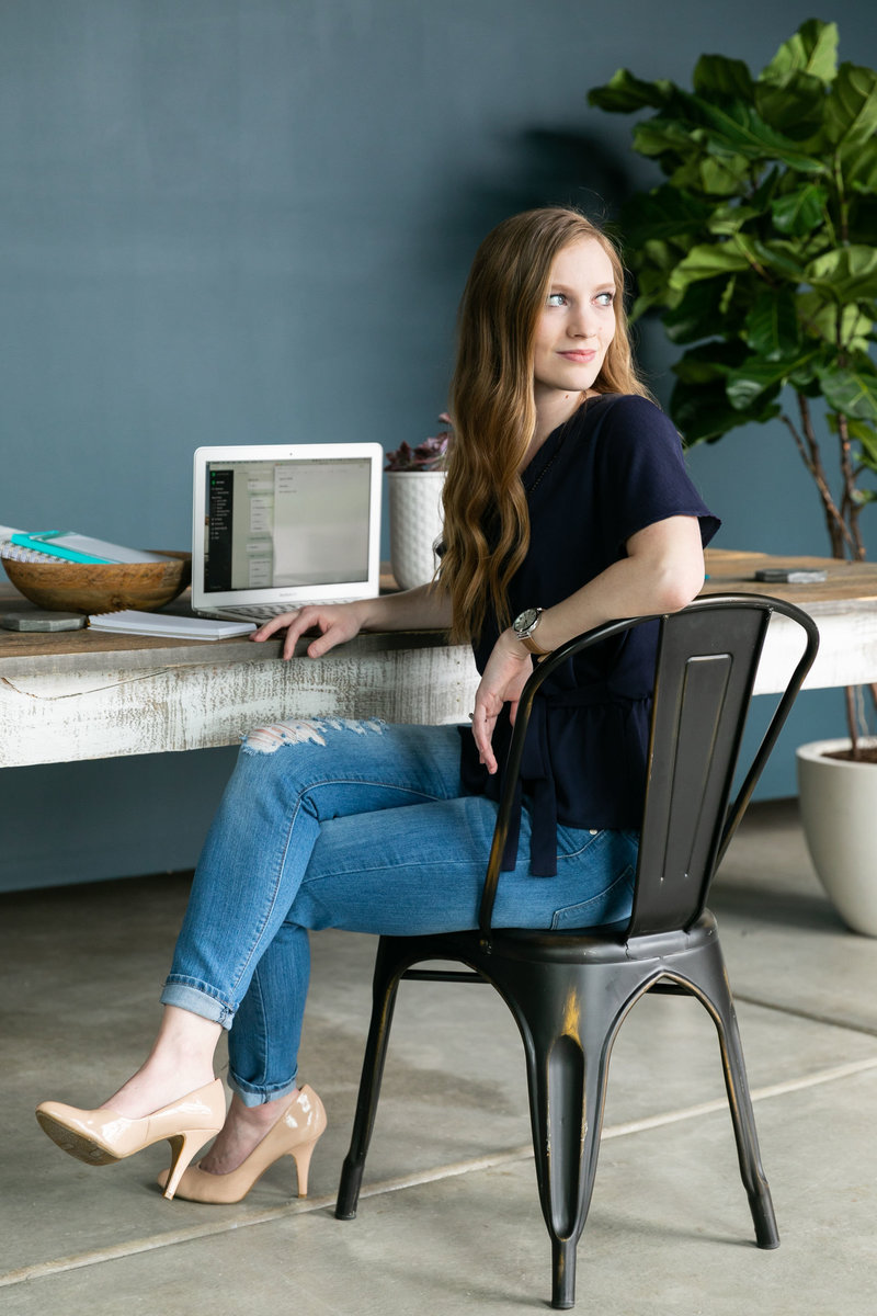 A woman wearing a navy shirt and ripped jeans sits behind a Mac computer