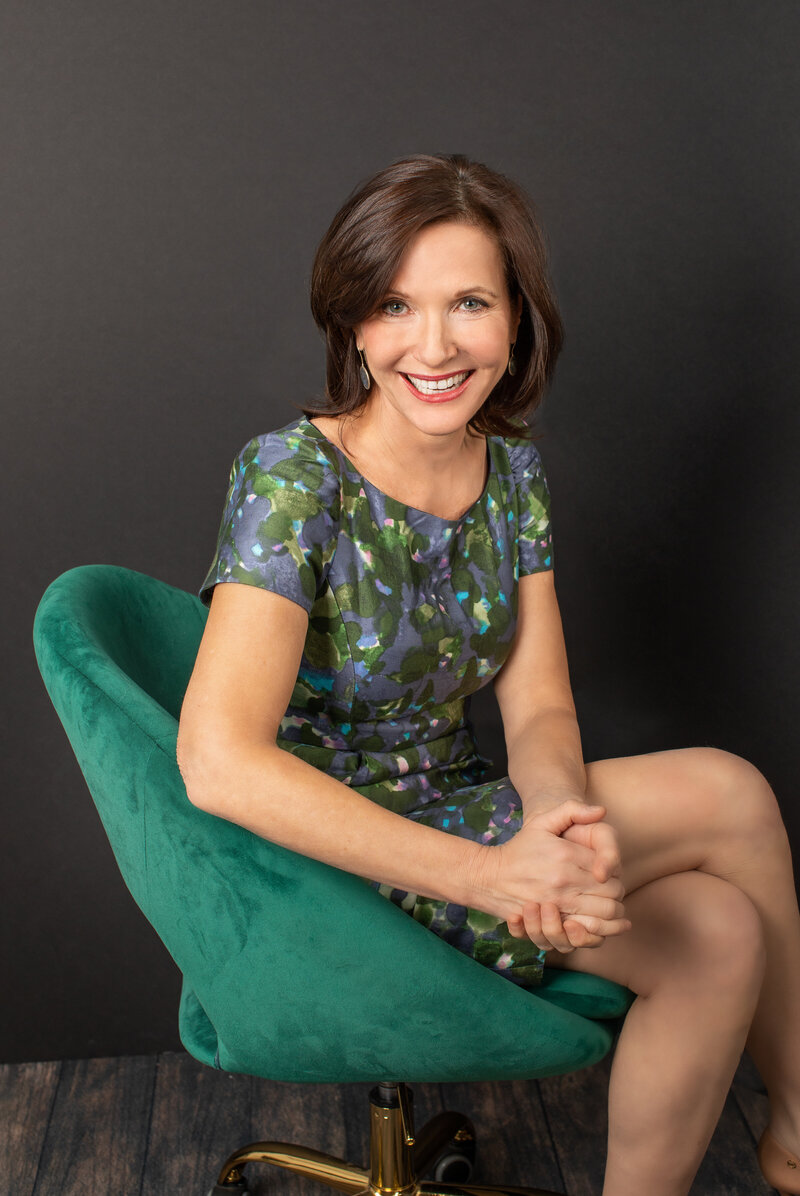 A woman wearing a green dress sits in a green chair smiling