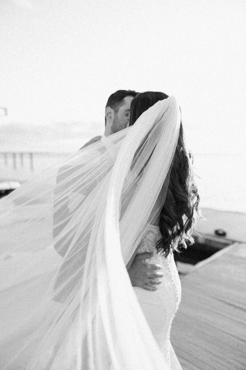 Black and white photo of a bride and groom embracing on a dock, with the bride's veil blowing in the wind, captured by a Luxury Wedding Photographer.