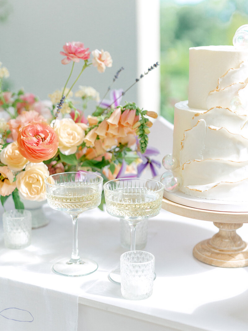 White tiered wedding cake on a stand next to a colorful floral arrangement and two glasses of champagne