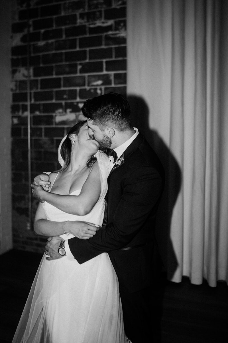 Newlyweds sharing a kiss, bride in a sleeveless white dress, with a brick wall background.