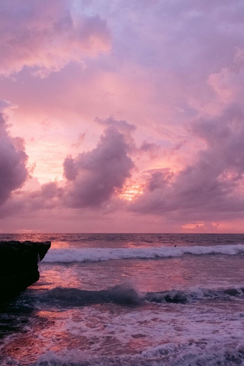 Pink and purple sunset over the ocean with waves crashing near a rocky shoreline.