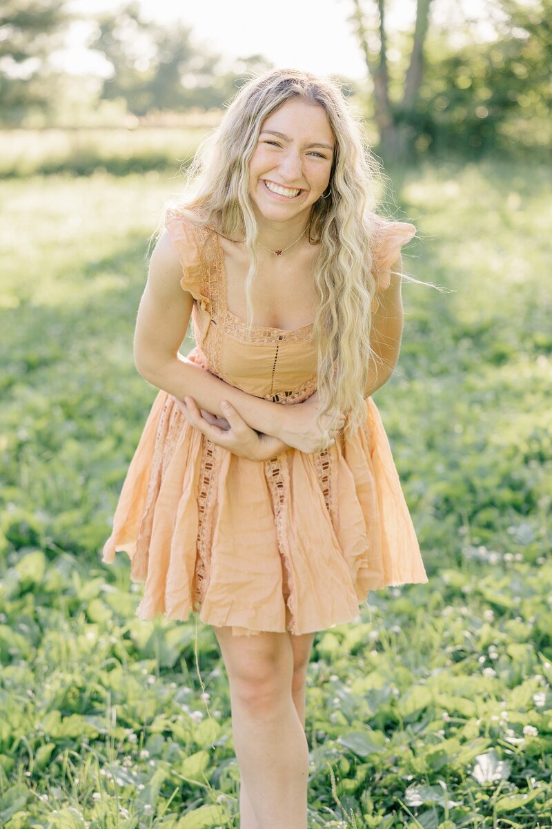 Senior girl in an orange free people dress laughing towards the camera in a green pasture, Indianapolis senior photographer