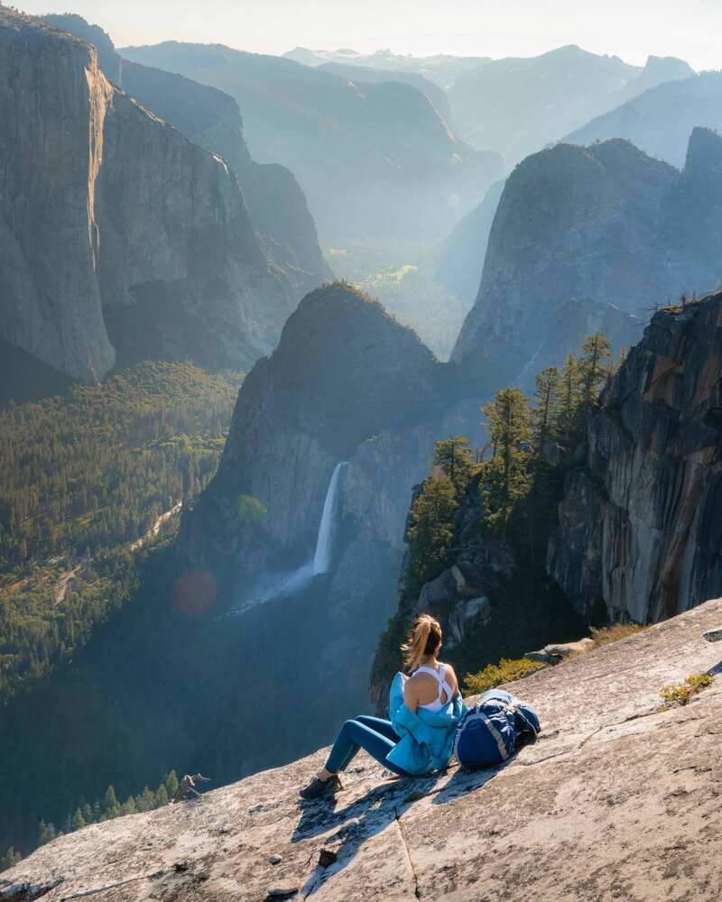 Woman in a blue jacket sitting on large rock looking out at a waterfall on a mountain range