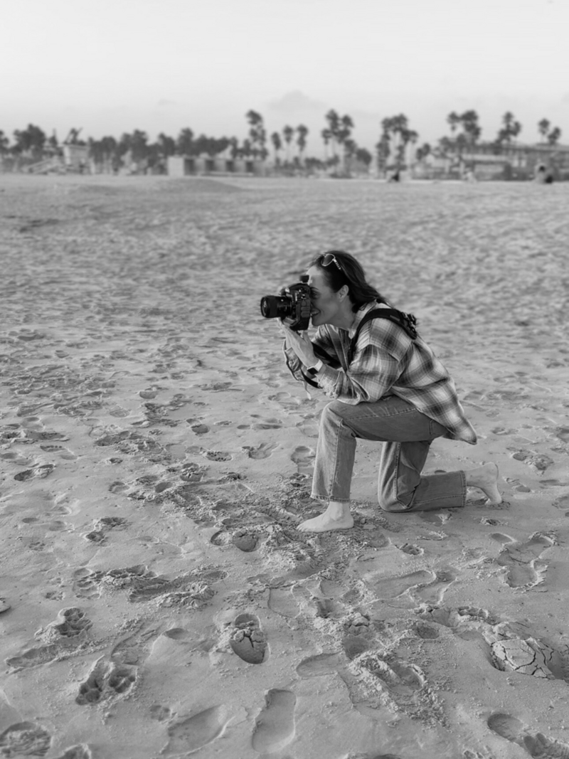 photography on beach in los angeles holding camera, kneeling on sand taking a photo wearing a plaid shirt and jeans black and white