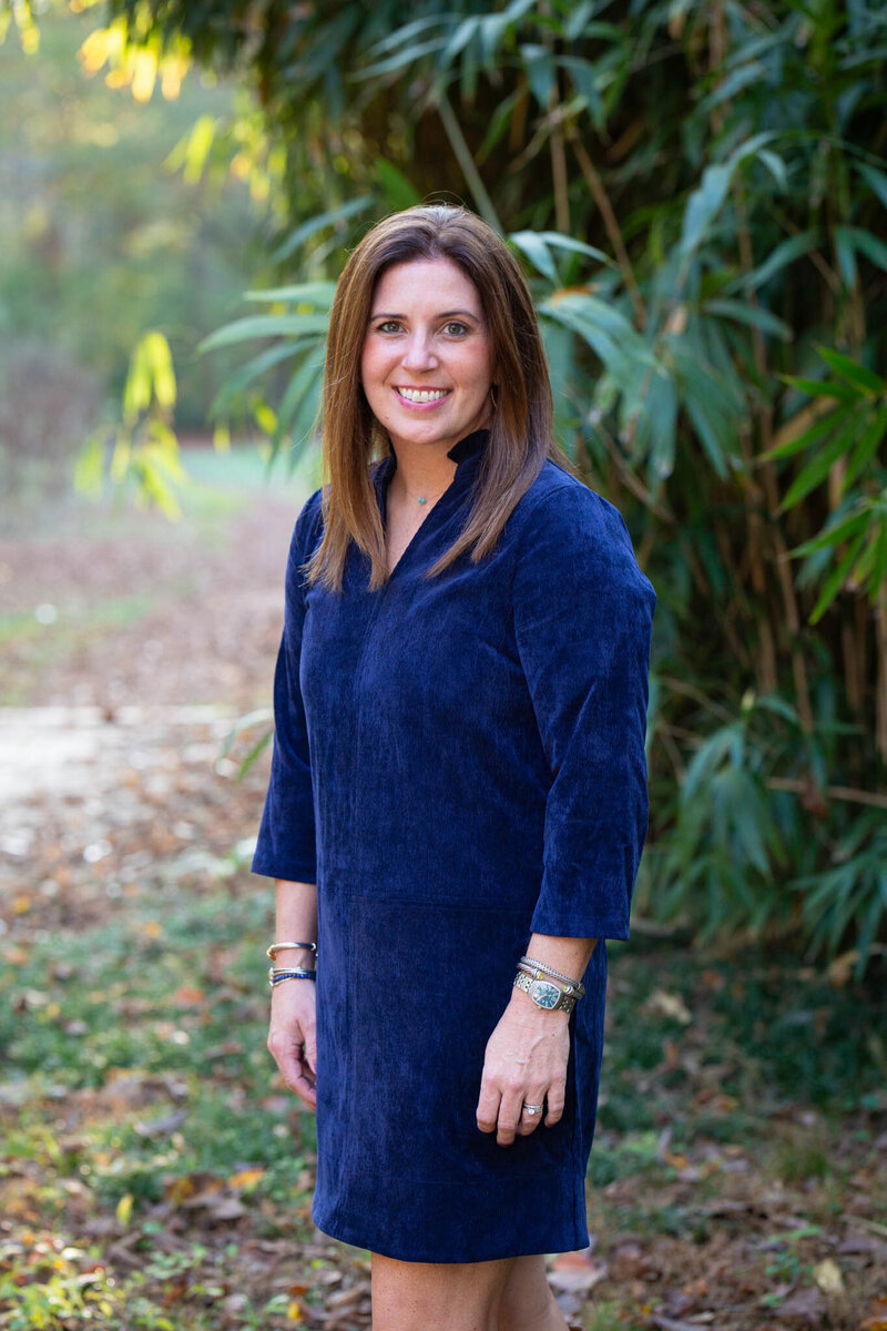 A woman in a blue dress stands outdoors, smiling. She is in front of tall green plants, with a blurred background of trees and fallen leaves, creating a natural, serene setting.